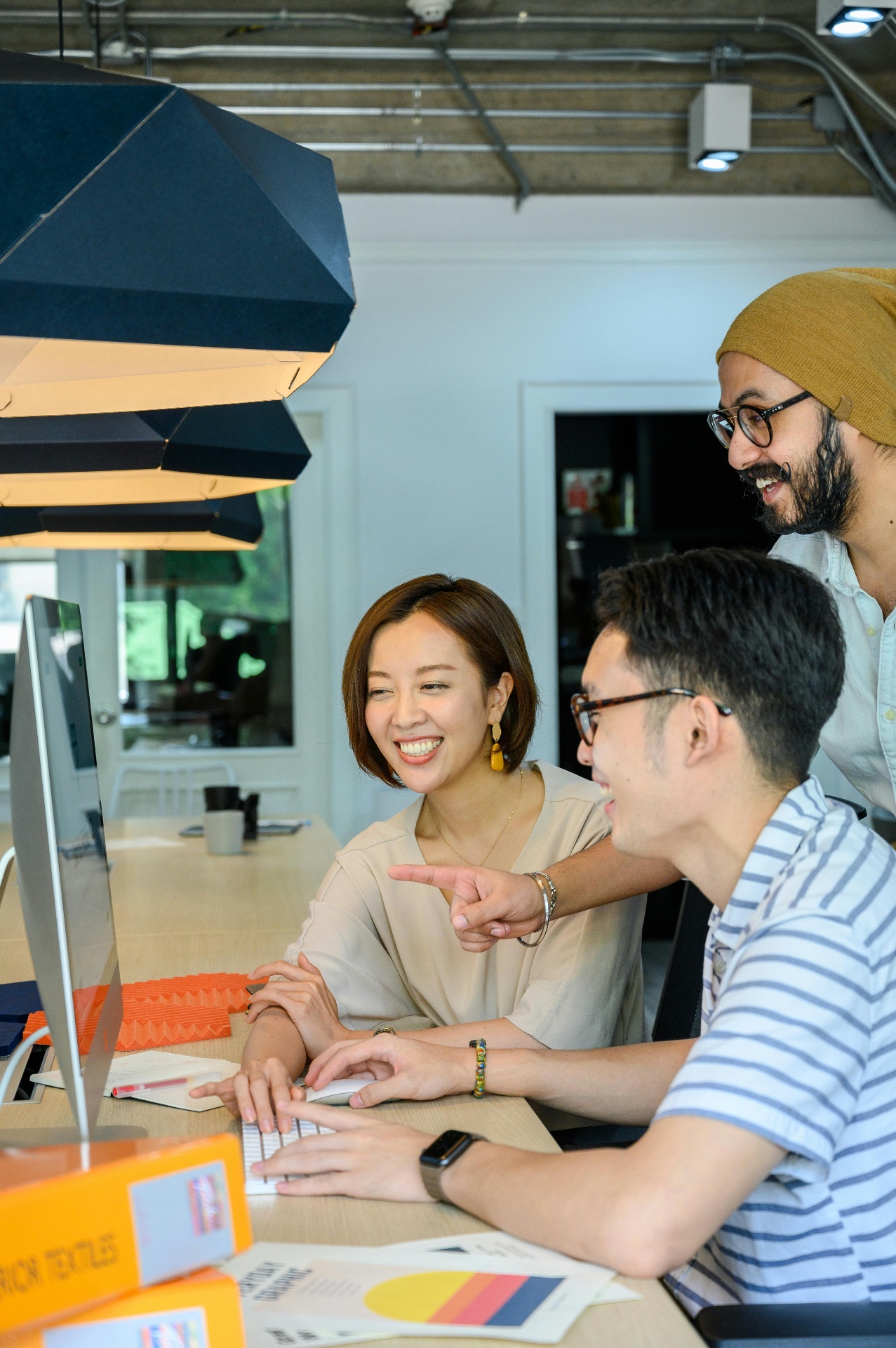 A communications team gathered around a large monitor in a warm office, pointing at a synthesis dashboard showing clusters of participant quotes grouped by theme with participant counts