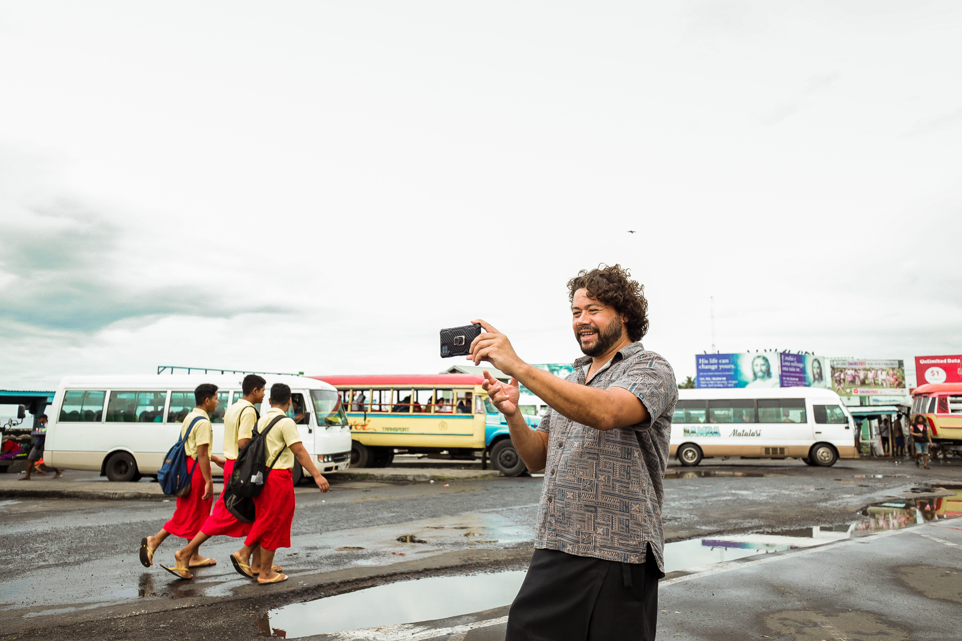 A man smiling while taking a photo on his phone in a busy street market